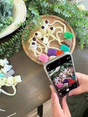 A top-down shot of a table setting featuring a wooden tray with various crocheted items and a phone capturing an image. A partially visible hand holds the phone, and there are decorative plants in the background.