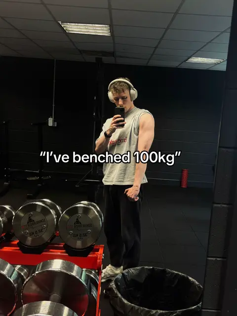 A man in a gym takes a mirror selfie, showing off his arm. He wears headphones and a grey t-shirt, and the text overlay says "I've benched 100kg". Behind him are weights and gym equipment.
