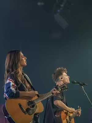 A live stage performance featuring two musicians playing acoustic guitars and singing into microphones. The image is primarily dark, with a spotlight illuminating the two performers. The woman to the left is holding a guitar and wearing a jacket with a British flag design. The man on the right is also holding a guitar and appears to be singing. The background is a gradient from dark to light, suggesting a concert or performance setting.