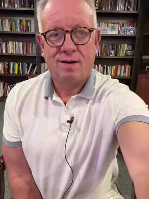 A medium shot shows a man with glasses, a white polo shirt, and a graying hair, sitting in front of a bookshelf.