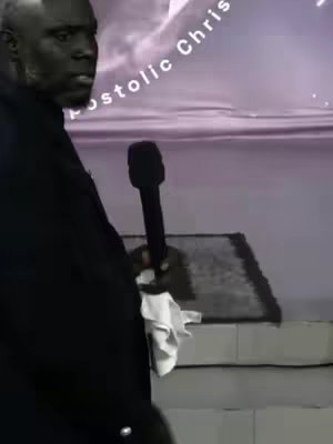 A man holding a microphone with text partially visible. The man is wearing a black suit, standing in front of a wall. The background is a blurred purple color. A cloth is visible over the stairs.