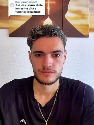 A young man with short, styled brown hair and a trimmed beard is centered in the frame. He wears a black t-shirt, a gold chain necklace with a religious pendant, and is looking down with a serious expression. Behind him is a wall with a painting featuring a gold cross and a blurred, abstract image.