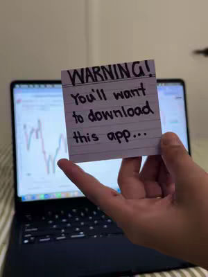 A person holds a sticky note up, obscuring part of a laptop screen that shows a financial chart. The note says "WARNING! You'll want to download this app..."