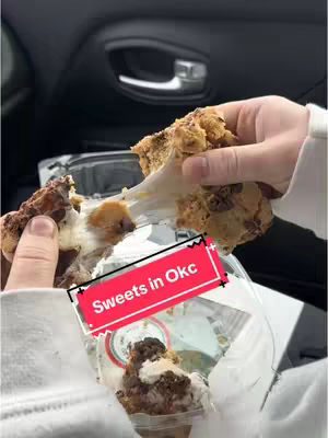 A first-person shot inside a car captures hands holding a partially torn cookie, with gooey filling stretching between the halves. Below, another cookie sits within a clear container. A text overlay reads 'Sweets in Okc' against a red background with a black outline, and the car interior setting is visible.