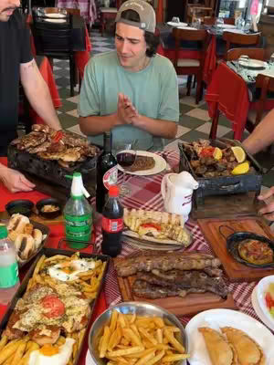 The image showcases a man seated at a table laden with various dishes, including grilled meats, fries, and sides. He's positioned in the center, wearing a neutral expression, with his hands clasped together in a gesture of anticipation or appreciation. The table is set with red and white checkered tablecloths, alongside bottles of Coca Cola and other drinks, suggesting a restaurant environment. The photo is lit with what looks like natural light and the variety of foods creates a visually appetising composition.
