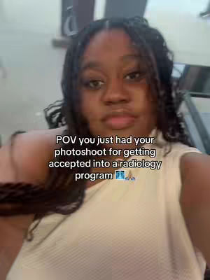 A selfie-style image featuring a young Black woman. She has dark hair styled in braids. Overlayed on the image is text that says 'POV you just had your photoshoot for getting accepted into a radiology program'. The composition is a medium shot with the woman near the center. The background is blurred, with elements appearing to be from an indoor setting. The color palette is natural, with the woman's skin tone and a cream-colored top being dominant.
