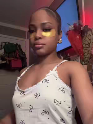 A woman with a light skin tone is looking directly at the camera. She is wearing gold eye patches under each eye, a white tank top with black bow prints, and gold hoop earrings. Her hair is pulled back from her face, and she is in a setting with a bright screen in the background. The lighting is somewhat soft.