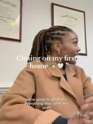 A woman with braided hair is captured in a medium shot, slightly turned to her right, smiling and looking off-camera. She's wearing a tan overcoat and a dark top. In the background, two framed documents hang on a light colored wall. The text overlay reads 'Closing on my first home' with a house and heart emoji.