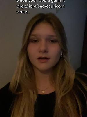 A young woman with long blonde hair is the central focus, positioned in a medium shot. She has fair skin and looks directly at the camera with a slightly serious expression. She's wearing a black top and a delicate necklace, and has earbuds in her ears. Text is overlaid at the top, and the background appears to be an interior space.