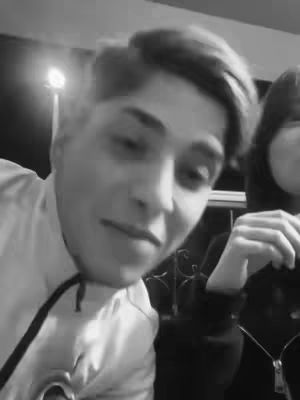 A black and white photo featuring a person close up, looking toward the camera. The person is wearing a light-colored collared shirt on a casual jacket; another person is partially visible next to them.