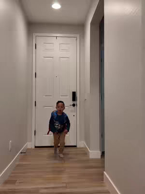 A young child wearing a backpack walks towards the camera, down a hallway. The hallway has light walls, wood floors, and a white door at the end. A bright light shines from a ceiling fixture.
