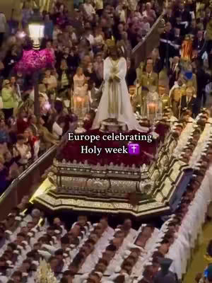 An overhead shot of a religious procession in a European city. The frame is filled with a crowd of people, many holding cell phones to record the event. In the center, a large platform is carried by a group. On top of the platform is a golden figure in white robes, likely a religious statue. The overall color scheme is a mix of earthy tones from the buildings, clothing, and the platform itself. Pink flowers add a contrasting pop of color near the top of the frame. Added text overlay provides context to the event, identifying it as Holy Week.