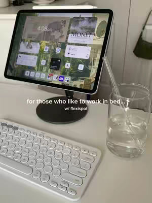 An iPad on a stand, with a keyboard in the foreground and a glass of water visible. The background is a clean, minimalist setup suggestive of a workspace.