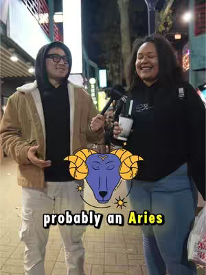 A street-level shot featuring two people, a man on the left and a woman on the right, both smiling. The man is wearing glasses, a black hoodie under a brown jacket, and holding his hand open to the side. The woman wears a black hoodie and is holding a microphone and a white cup. Between them is a graphic of a zodiac ram symbol. The background shows a city street.
