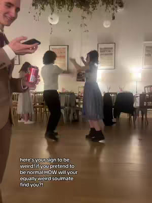 In a well-lit room, a person holding a phone and a can are in the foreground. Two individuals are dancing in the background, while the image contains text overlayed.