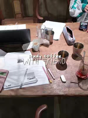 A tabletop scene, dominated by a light-brown wooden surface, is littered with study materials and coffee-related items. Several stainless steel tumblers sit among scattered papers with writing and printed images, a pen, a pencil, and an eraser that suggest note-taking and studying. A phone is visible. The words 'study coffee WITH FRIEND' are overlaid in a handwritten-style font. The overall composition evokes a casual, communal study session.