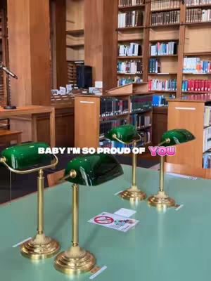 The image shows an interior of a library with wooden bookshelves filled with books. On a light green table in the foreground are four green-shaded lamps with brass bases. Text overlays on the lamps.