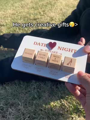 A person is holding a white wooden sign that says "DATE NIGHT" with cubes and a small red heart in a grass field, the sign has cubes with date ideas written on them such as "GO FOR A WALK" and "WATCH A MOVIE". In the background you can see the person's black shirt, and hands in the photo holding the sign.
