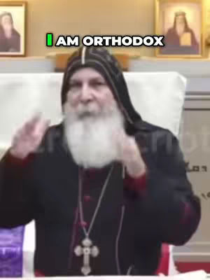 A man in a black robe with a white beard is speaking, with the text overlay, "I AM ORTHODOX", above him. In the background are two framed pictures.