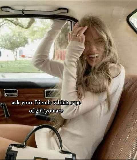 A woman is sitting in the passenger seat of a vintage car, laughing with her hand up to her head. The interior of the car is visible, with a tan seat and a wooden door panel. Outside, a blurred green background hints at a park-like setting.