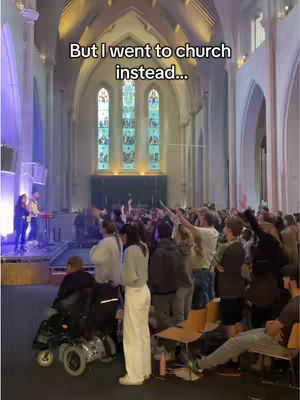 A wide shot inside a church shows people gathered for a service. The focus is on the crowd, with some raising their hands in what seems like worship. The architecture of the church is prominent, with tall arches, stained-glass windows, and a stage area. The lighting is a mix of natural light from the windows and artificial stage lighting.
