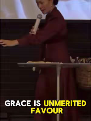 A woman in a burgundy dress stands at a podium, holding a microphone. Text overlays at the bottom read 'GRACE IS UNMERITED FAVOUR' in white and yellow.