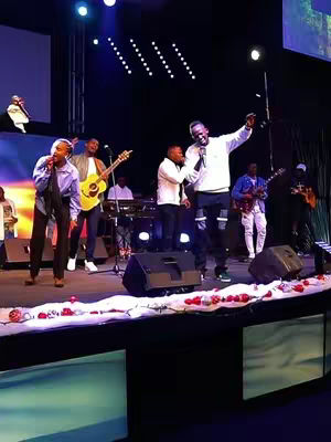 The cover image captures a live performance on a stage, with a group of musicians and singers. The stage is well-lit, and a backdrop with a screen is visible. A lead singer is holding a microphone and addressing the audience, with several other performers visible. The color scheme is warm and the lighting emphasizes the performers. The layout indicates a live music setup.