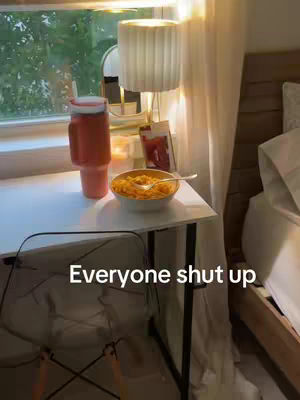 A cozy indoor scene at a table next to a bed. A table holds a bowl of food, a drink with a straw, and other objects in soft lighting. The text overlay reads: Everyone shut up.