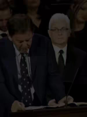 The cover image features two men in suits seated indoors. The man on the left is speaking, slightly looking down and to his left. The man on the right, wearing glasses and a black tie, is looking forward with a neutral expression. The background is out of focus and primarily dark, suggesting an audience or event setting. The lighting is subdued, with focused light on the subjects.