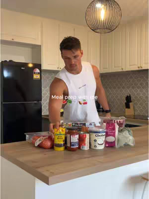 A man stands in a modern kitchen, preparing what appears to be meal prep. Various food items are arranged on a countertop, and the overall lighting is bright.