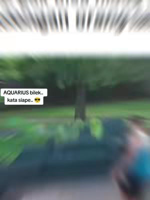 The image is a blurry outdoor scene. The background appears to be a green area with trees and foliage. In the foreground, there's a slightly out-of-focus area, which could contain people or other details. Overlayed on the image is a text box with the text 'AQUARIUS bilek.. kata siapa...'. The overall effect is a mix of natural elements with superimposed text, hinting at an astrological theme. The central focus is the superimposed text.