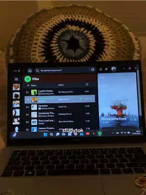 A laptop displaying a Spotify playlist with a blurred-out, textured background and a crocheted circular object visible behind the laptop. The laptop screen shows a Spotify interface with a playlist titled "Vibe." The overall composition is centered on the laptop, with the background elements adding a soft, textured visual interest.