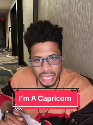A man with glasses is centered in the frame, speaking, with a red banner overlayed across the bottom that says "I'm A Capricorn." The background is a blurred hotel room or similar interior.