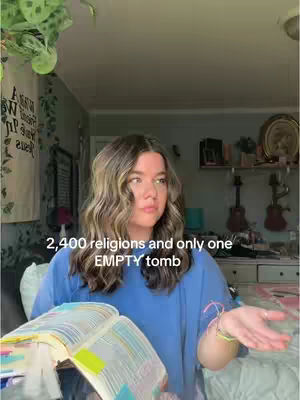 A young woman with long, wavy brown hair is sitting indoors, holding a large book with colorful sticky notes. She's wearing a blue shirt and has bracelets on her wrist. Her room has a light color scheme, and behind her are decorations including a framed print and some wall decor. Overlaid on this image is text that reads '2,400 religions and only one EMPTY tomb'.