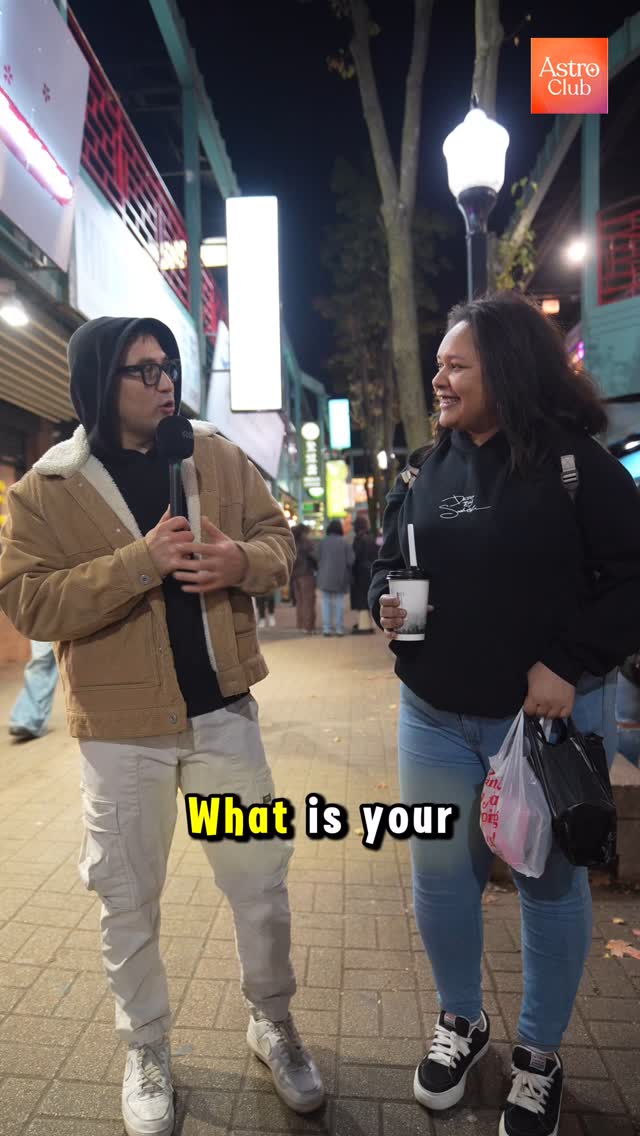 A man holding a microphone interviews a woman on a city street at night. Overlaid text asks a question. In the background, a street with shops and other people are visible.