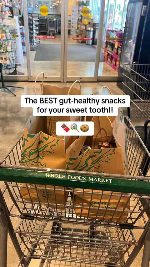 The cover image shows a shopping cart filled with two brown paper Whole Foods Market bags. In the bags, various food items are visible. A text overlay reads "The BEST gut-healthy snacks for your sweet tooth!!" with emojis of a chocolate bar, a lollipop, and a cookie.