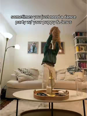 The image shows a person holding up a small dog in a living room. The person is wearing casual clothing and standing in front of a white couch and a coffee table. There's text overlaying the image.