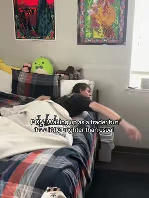 A person is lying in bed with a patterned comforter and a white sweatshirt. A green plush toy and two framed artworks are on the wall behind the bed. The text overlay says: 'POV: Waking up as a trader but it's a little brighter than usual'.