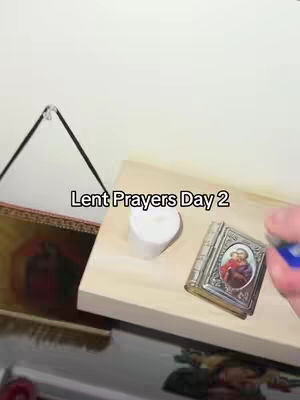 The image displays a small display on a white wall. On a wooden surface, there's a white candle, a small book with a religious image, and a hand holding a lighter. A framed picture hangs on the left. The text overlay reads 'Lent Prayers Day 2.'