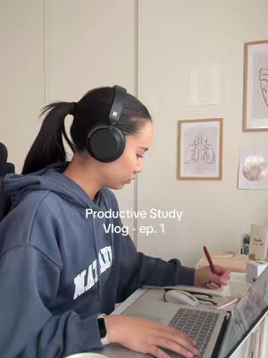 A woman is seated at a desk, facing slightly left, wearing headphones and a blue hoodie. She's working on a laptop, with a pen in her right hand, appearing to take notes. The background includes framed artwork and a wall, creating a study environment. The image has a soft lighting.