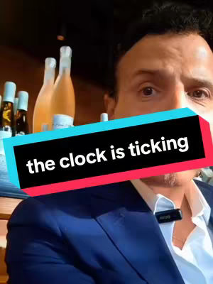 A man in a blue suit and a white shirt is looking directly at the camera. A rectangular text overlay in a black background with red border emphasizes the phrase “the clock is ticking.” Behind him, bottles and glassware indicates an elegant location.
