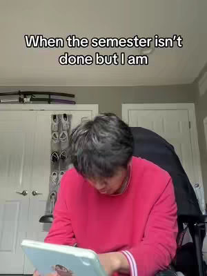The image shows a young man in a pink sweater looking down at a tablet, appearing to be studying. Above him, text reads 'When the semester isn't done but I am'. The background shows a room with a white door, a closet with a few shoes hanging, and a closed door on the right. The lighting is soft, with the focus on the student and his task.