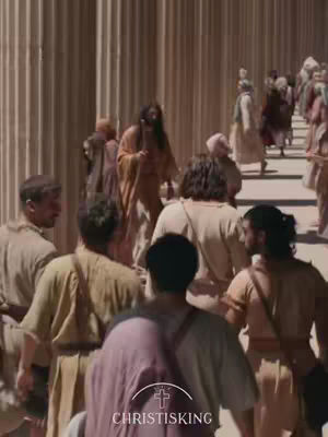 A scene from a historical film featuring numerous people walking towards the viewer. Tall, vertical columns form the background; the foreground shows a crowd of people dressed in ancient attire, with one central figure in an orange robe.