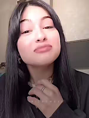 A selfie-style shot of a young woman with dark hair and fair skin. She's looking towards the camera with a slight, somewhat mischievous expression. Her lips are slightly parted. Long, dark hair frames her face, and a black top. A ring is visible on her finger. The background is neutral, keeping the focus on the woman.