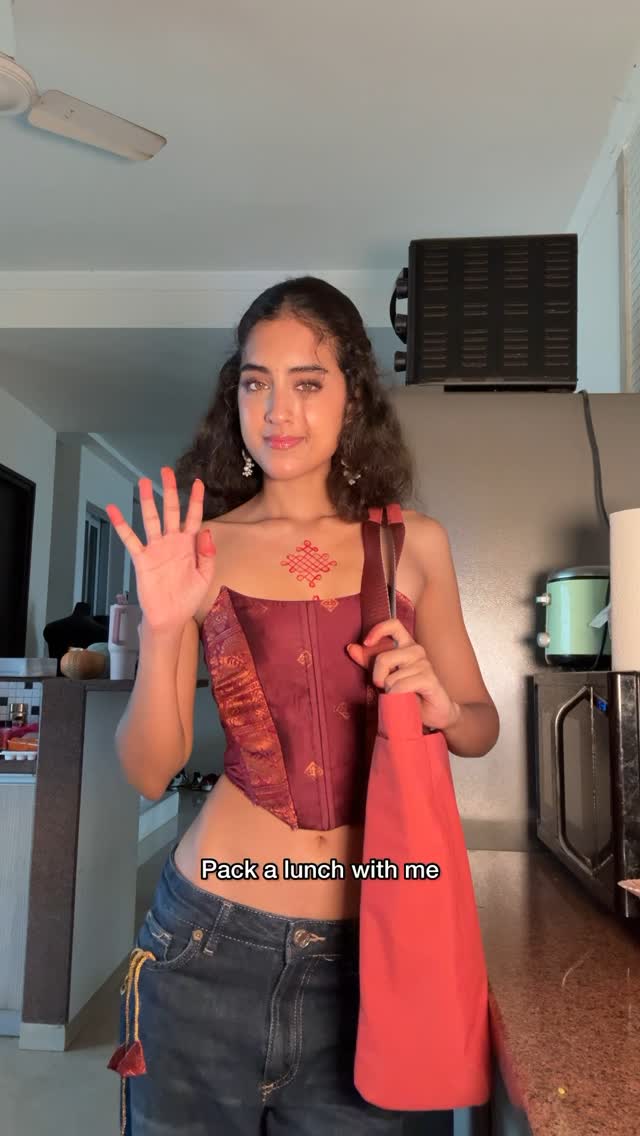 A woman in a maroon corset top and low-rise jeans stands in a kitchen, holding a red tote bag and gesturing with her right hand. The background includes kitchen appliances like a microwave and a cabinet.