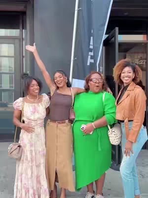 Four women are standing together smiling, posing in front of a dark building entrance. The woman on the left is wearing a pink floral dress. The next woman is wearing a brown shirt and long skirt, with her arm raised. The third woman is wearing a green dress. The woman on the right is wearing a brown jacket. In the background is a black flag.