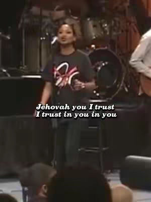 A woman is singing on a stage with a band in the background. The foreground features a dark set, some people and chairs. Text overlay is displayed at the bottom of image.