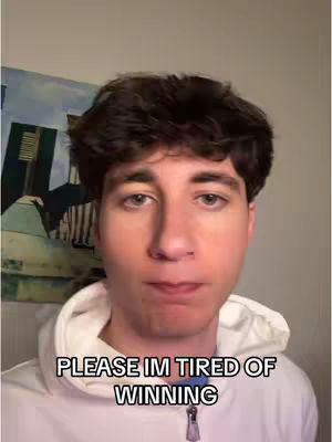 A young man in a white hoodie with dark hair looks directly at the camera. Text overlaid on the bottom of the image reads 'PLEASE IM TIRED OF WINNING.' The background is simple with muted colors.
