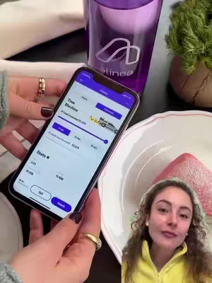 A woman holds a phone displaying the Alinea Invest app, set against a background that includes a water glass with the Alinea logo, a small plant, and a plate with a pink pastry. The woman is smiling and visible partially from the waist up, her face in the corner.