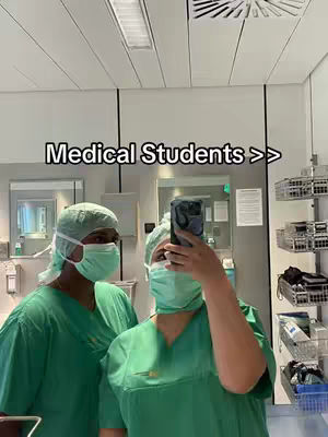 Two people in medical scrubs and surgical caps pose for a selfie in what appears to be a hospital setting. The person in front is holding a phone, obscuring their face. The person on the left is also wearing a mask and cap.